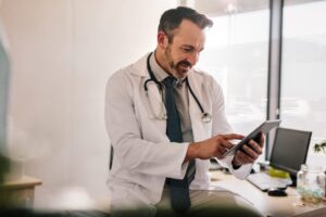 Smiling male doctor in white coat with stethoscope sits in office, using a tablet to review patient notes