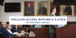 Attorney speaks at courtroom podium; colleagues listen, portraits behind, Williams Elleby Howard & Easter logo