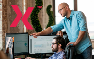 Manager points at analytics on dual monitors while colleague works; large pink X logo on exposed-brick wall