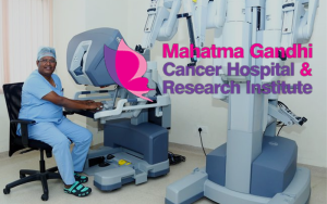 Surgeon at robotic surgery console smiles in scrubs, beside robotic arms, Mahatma Gandhi Cancer Hospital logo