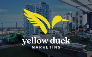 Yellow Duck Marketing logo with a stylized yellow duck in flight, set against a blurred modern cityscape background