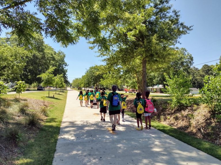 Children with colorful backpacks walk along a shaded park trail on a sunny day, guided by an adult leader